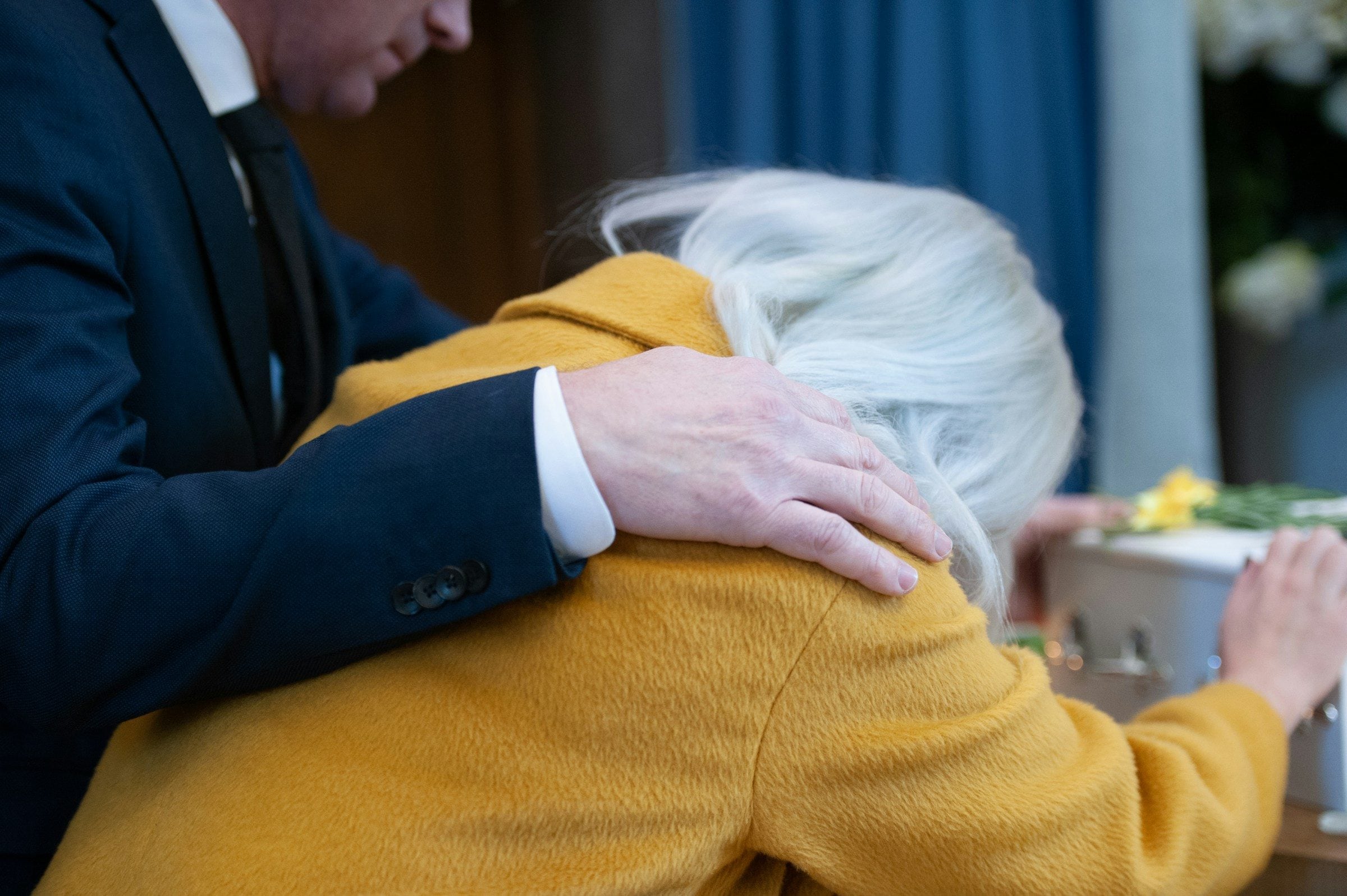 A hand resting on the shoulder of an older woman in a yellow cardigan
