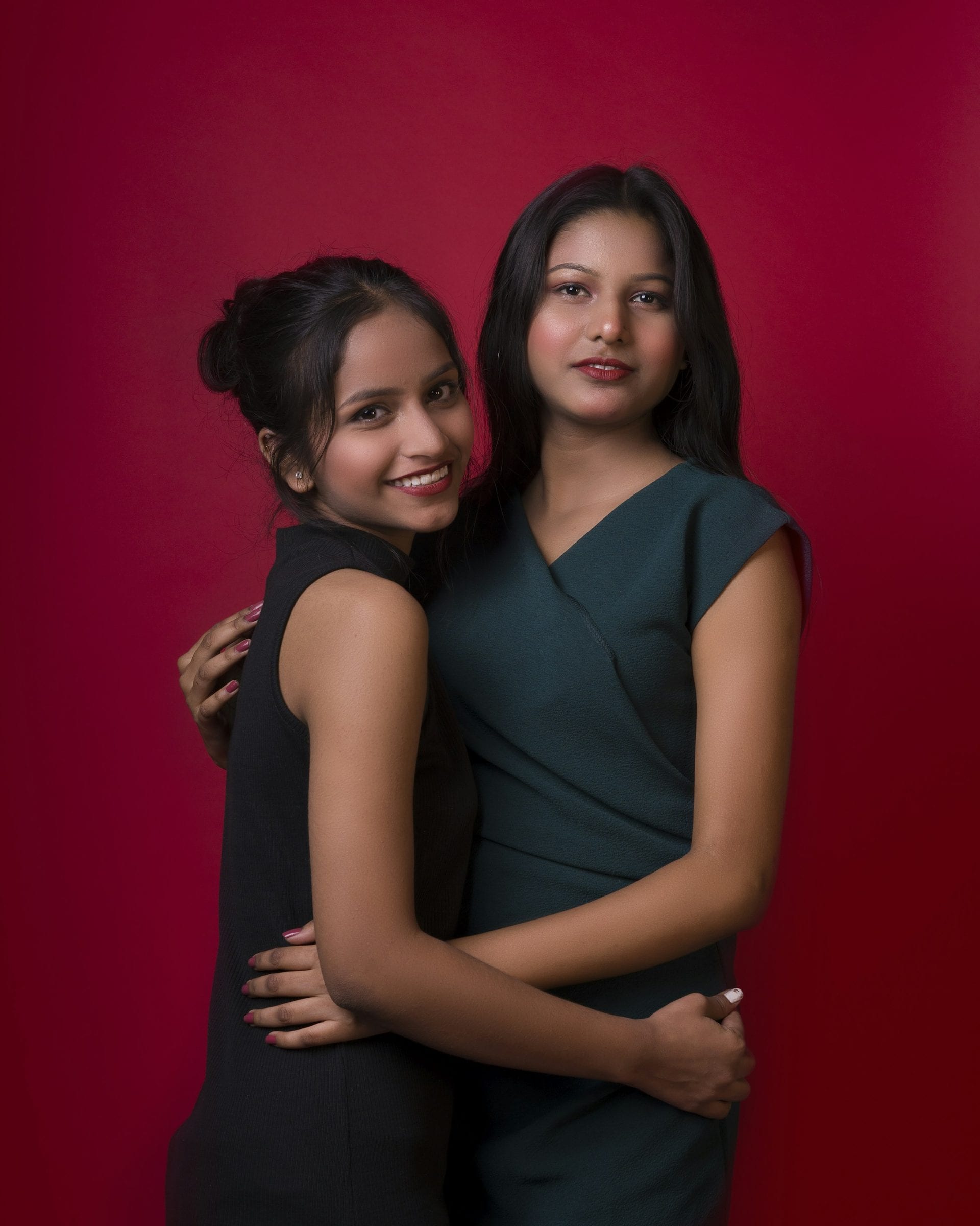 Two sisters holding each other, photographed against a deep red backdrop